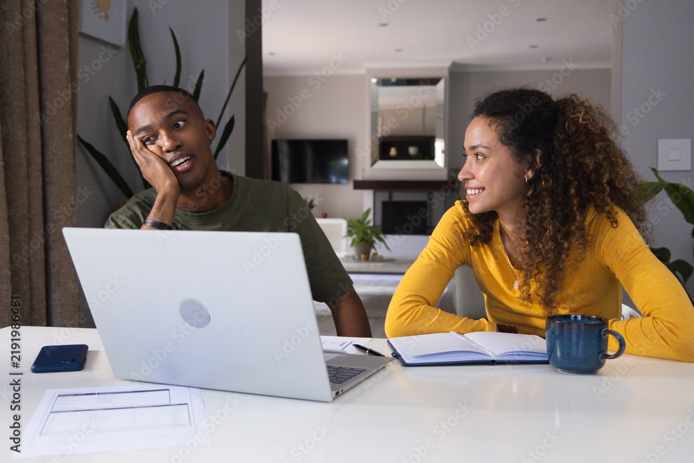 Obraz premium Couple sitting at open-plan living room table, sorting papers with laptop, mustard top, olive tee