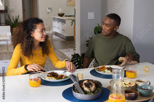 Diverse couple wearing yellow top and olive tee sitting at dining table sharing chicken and juice