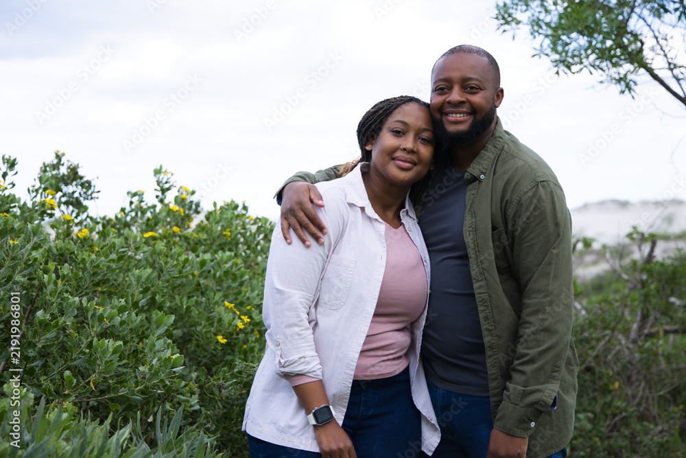 Obraz premium African couple standing among coastal shrubs near sandy dunes, wearing jeans jacket and smartwatch