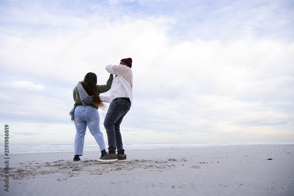 Obraz premium African American couple walking on wide sandy beach at shoreline, wearing jackets and beanie
