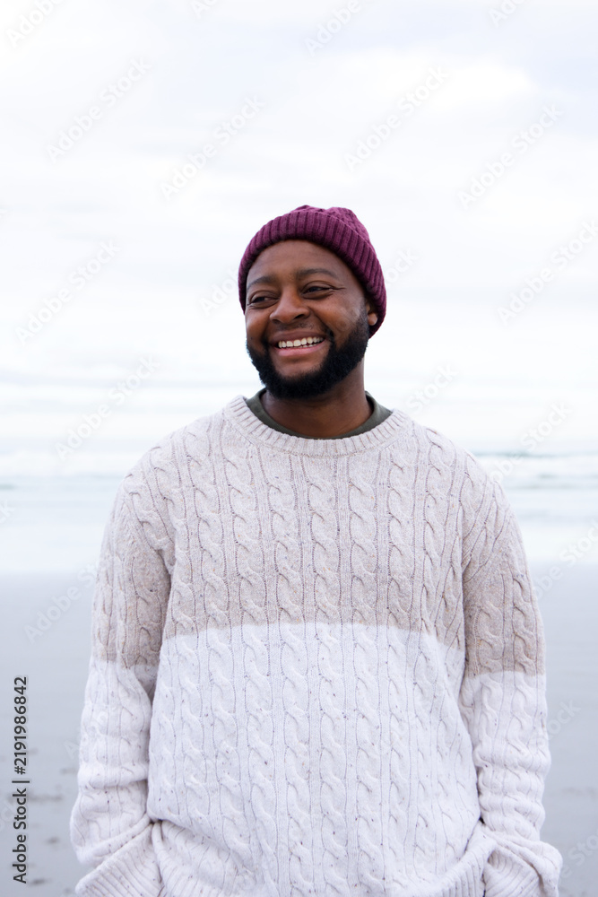 Obraz premium African man standing on wet sand shore, wearing maroon beanie and cream cable knit sweater