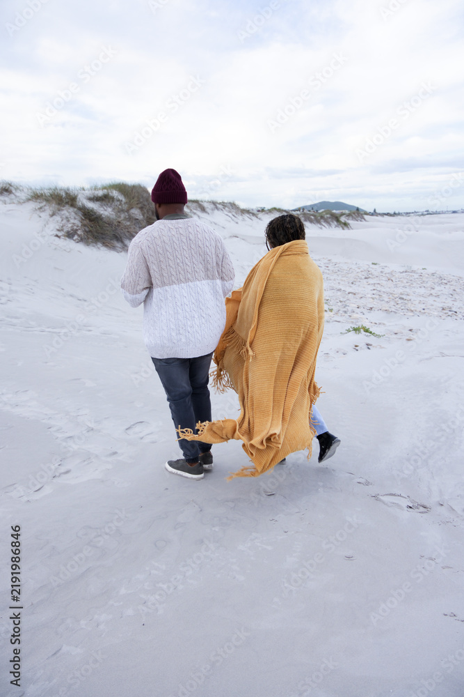 Obraz premium Couple walking across pale sand dunes at beach, showing mustard blanket, beanie, sweater