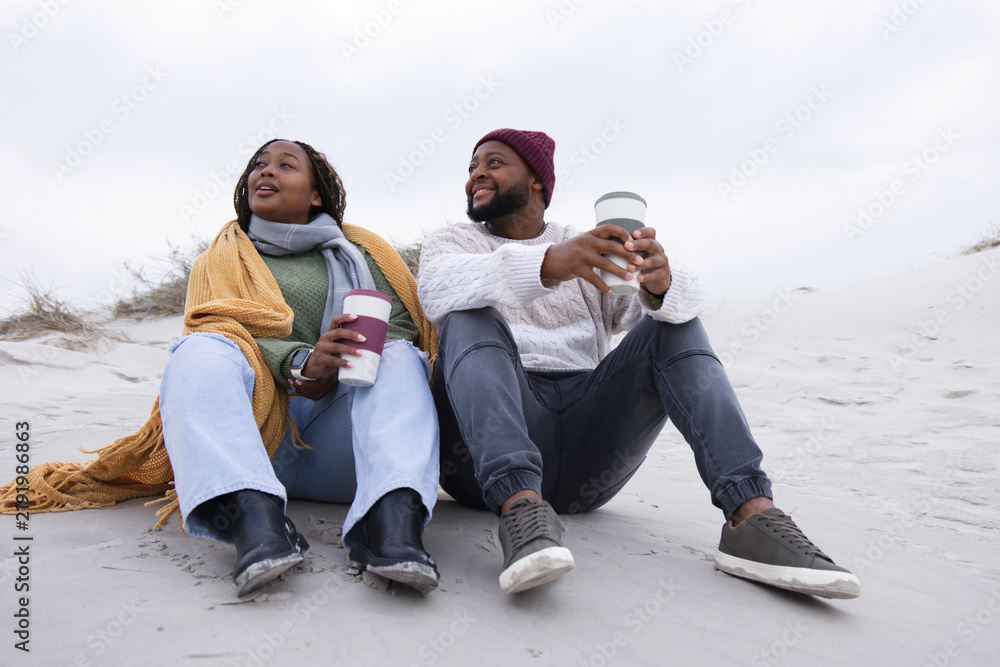 Obraz premium African American couple sitting on sandy dune at beach holding reusable travel cups, wearing shawl