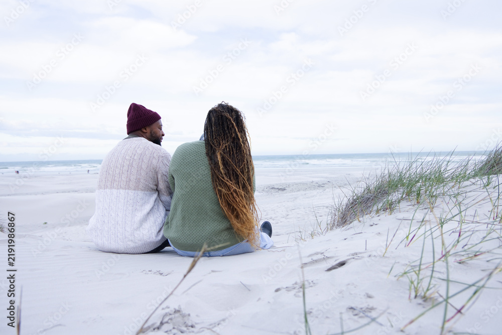 Obraz premium Couple sitting on sand dune at beach, wearing knit sweaters and beanie, showing footprints