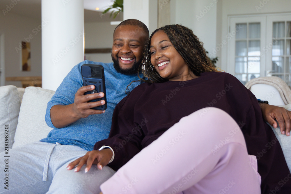 Obraz premium African American couple sitting on light sofa in living room sharing smartphone with bracelet