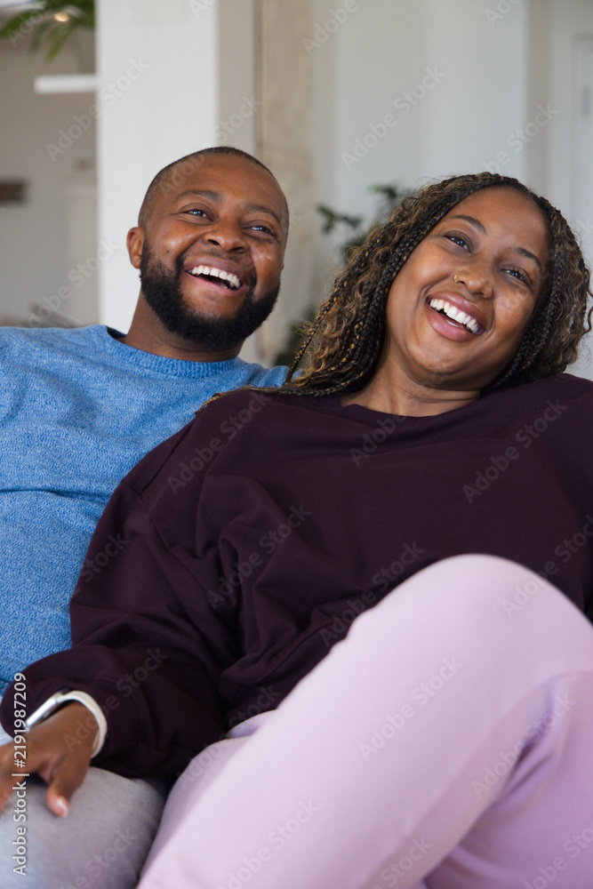 Fototapeta premium African American couple laughing, leaning on couch at home, woman in maroon top, white smartwatch