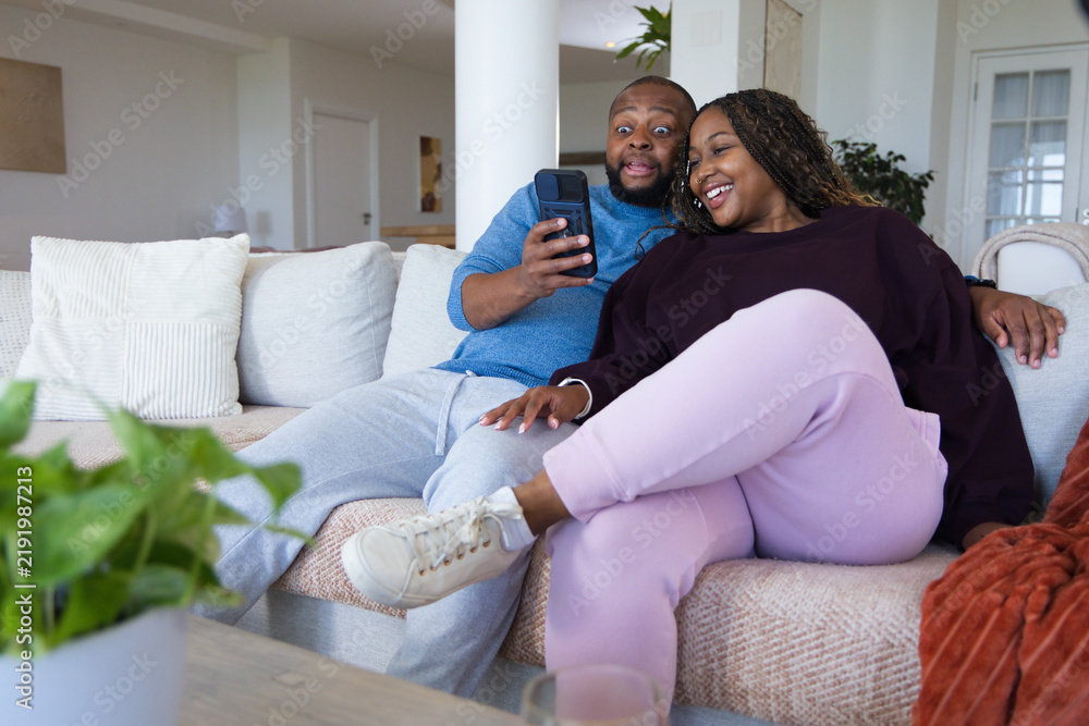 Obraz premium African couple sitting on light sectional sofa at home, man holding smartphone, woman smiling