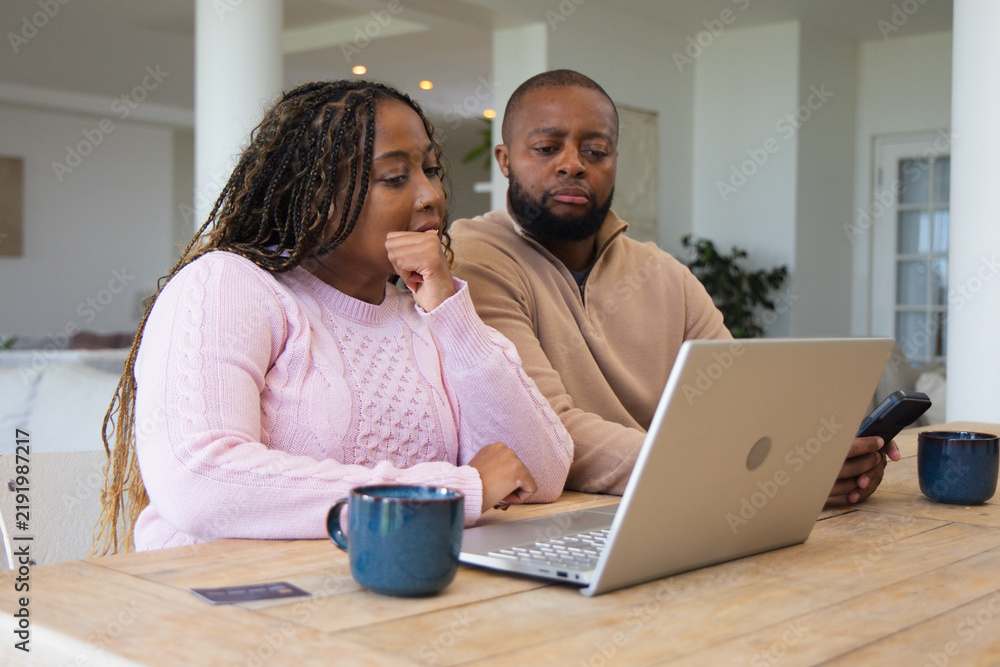 Obraz premium Couple sitting at wooden table at home, reviewing laptop with smartphone and woman in pink sweater