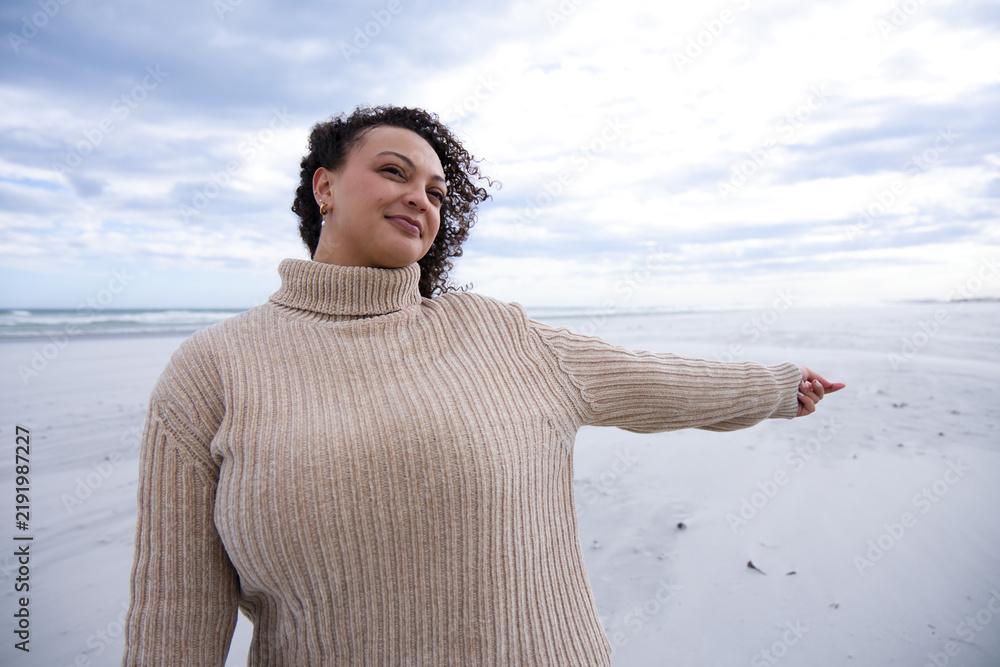 Obraz premium Adult African American woman pointing on beach wearing beige sweater hoop earrings ocean cloudy sky