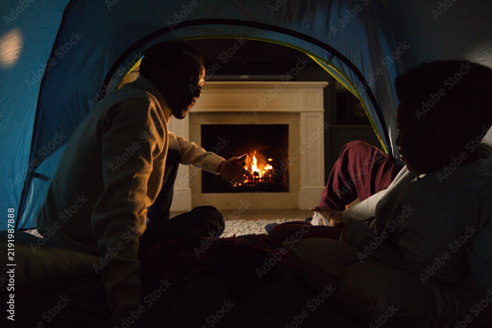 Obraz premium African American adult and youth males sitting in pop-up tent on living room rug facing fireplace