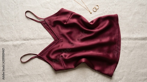 Deep red satin camisole top with delicate gold necklace on a textured linen background.