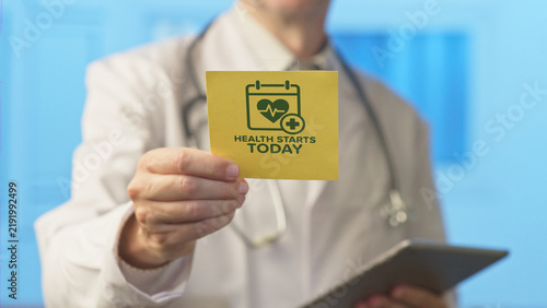 Doctor in a white lab coat and stethoscope holding a yellow reminder card with a calendar and heart icon, featuring the text health starts today emphasizing wellness and proactive healthcare