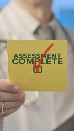 Person is holding a yellow sticky note showing the words assessment complete with a red checkmark confirming successful task accomplishment, representing evaluation results and achievement