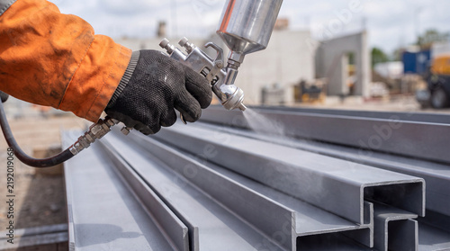 Worker sprays paint on steel beams at a construction site in daylight