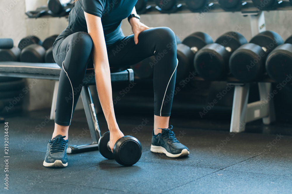 Obraz premium Woman lifting dumbbell in gym during strength training workout with focused energy, seated on bench beside rack of weights and exercising for fitness and muscle development