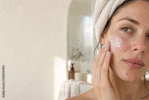 Woman with a towel turban smooths cream onto her cheek after a bath, evening skincare routine with copy space