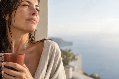 Woman with wet hair enjoys tea on a sunny balcony letting her hair air dry, slow self care at home with copy space
