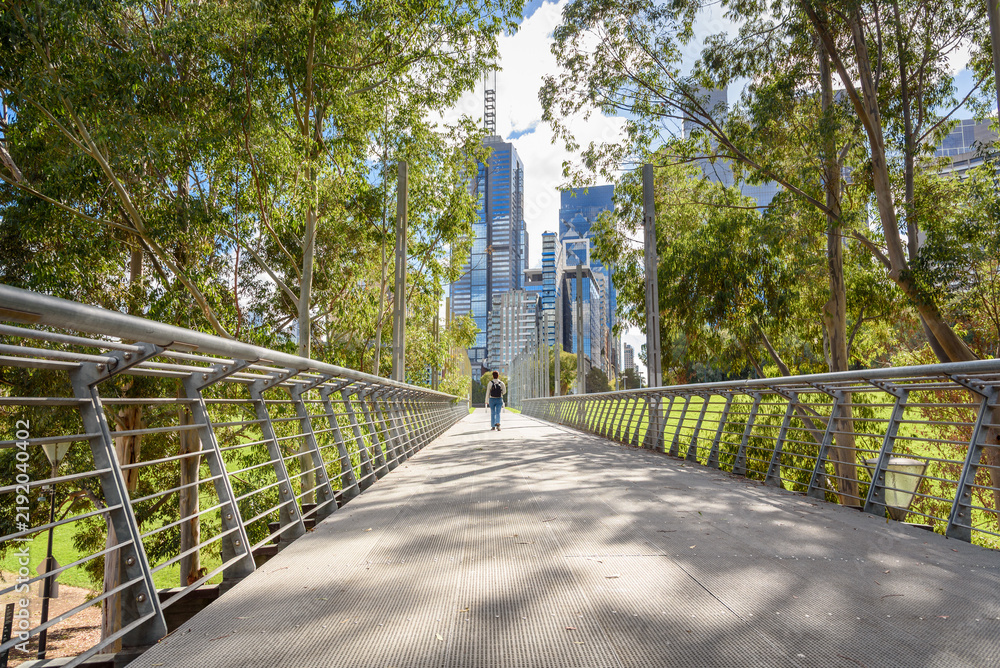 Obraz premium Woman walking on an elevated walkway lined with trees in a park bordered by modern glass office skyscrapers in a city centre on a sunny autumn day