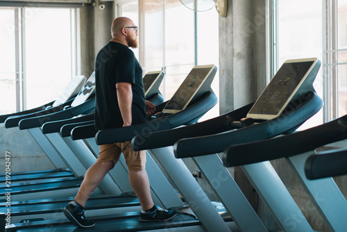 Obraz na plátně Bald man walking on treadmill in modern gym beside large windows with focused ex
