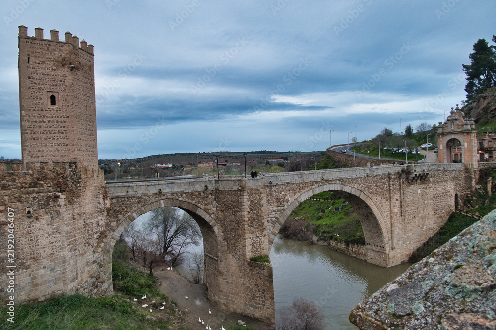 Obraz premium Historic Bridge and Tower Over Tagus Riv - Panoramic view of the iconic Puente de San Martín, a medieval arch bridge, and its fortified tower spanning the Tagus River in Toledo, Spain