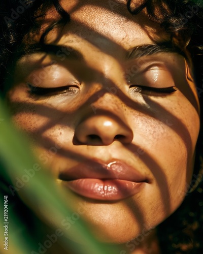 Close up portrait of woman with eyes closed in warm sunlight with hard shadow pattern from leaf on face. Natural light and tranquility concept. Summer serenity and mindfulness for wellness and beauty