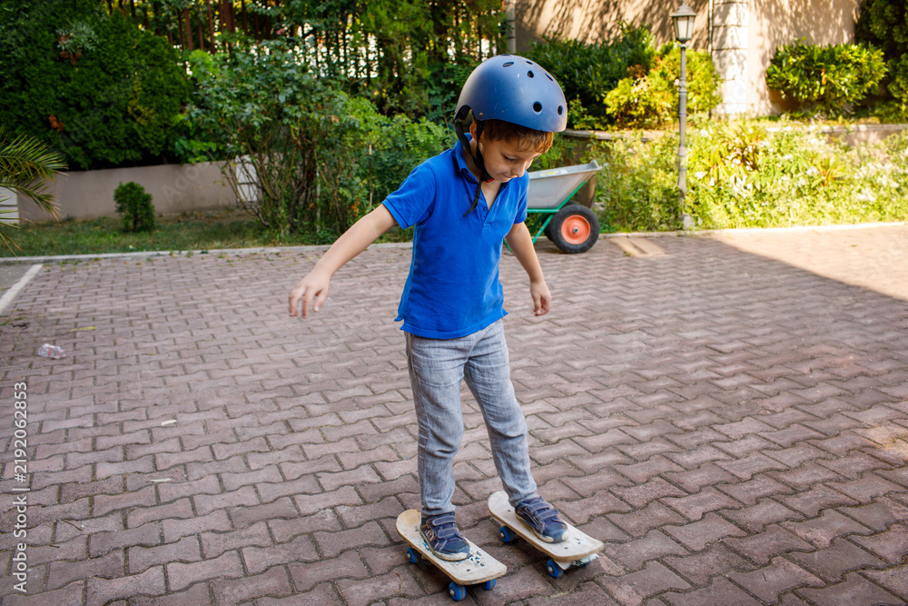 Fototapeta premium Young boy wearing safety helmet skateboarding in backyard, brick paved surface, garden with potted plants and wheelbarrow, residential buildings in background, active childhood outdoor recreation