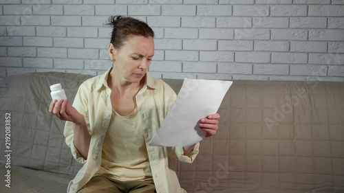 Confused mature woman reading medical prescription. Puzzled mature woman sitting on a sofa while reading the instructions on a medicine bottle and a prescription, showing a bewildered