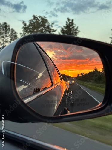 Canvas Print Close-up of a car side mirror reflecting a fiery orange and cloud-filled sunset sky while driving down a countryside road