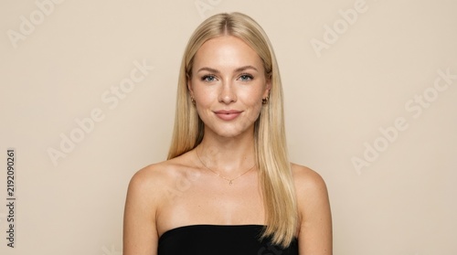 Young blonde woman facing camera in a black strapless top, clean beauty studio portrait, pale neutral background, even soft light, glossy straight hair, realistic skin texture, symmetrical composition