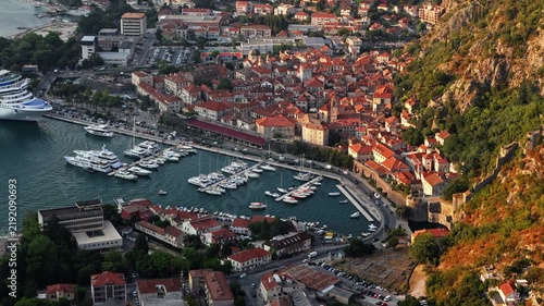 aerial drift across a mountain slope in low evening sun with warm light on the red roofs and stone walls of kotor old town and dark calm water in the marina, montenegro