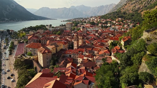 long aerial flight from a wide view of kotor down to the twin stone bell towers of the old town church, with a busy square, red roofs and the hazy bay behind, montenegro