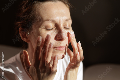 Fotografie Mature Woman Applying Face Moisturizer in Warm Morning Sunlight
