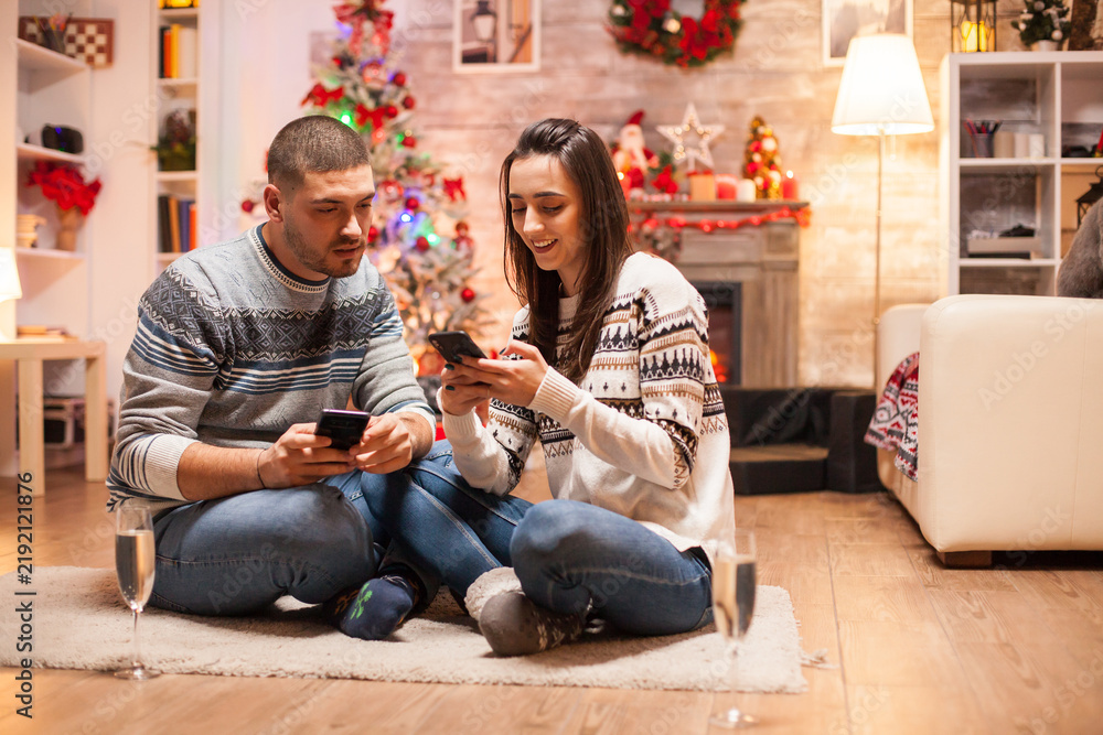 Obraz premium Couple sits close together on the floor using separate smartphones beside a decorated tree and fireplace during relaxed Christmas downtime at home.