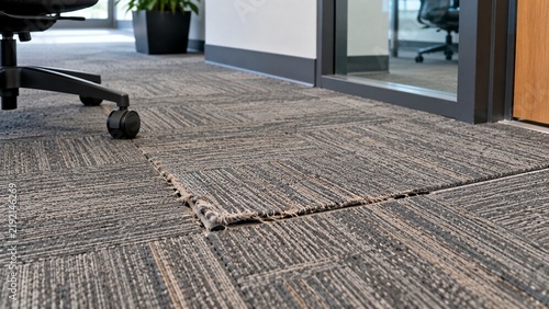 Close-up of commercial office floor showing one badly worn carpet tile with frayed edges and visible separation from adjacent tiles
