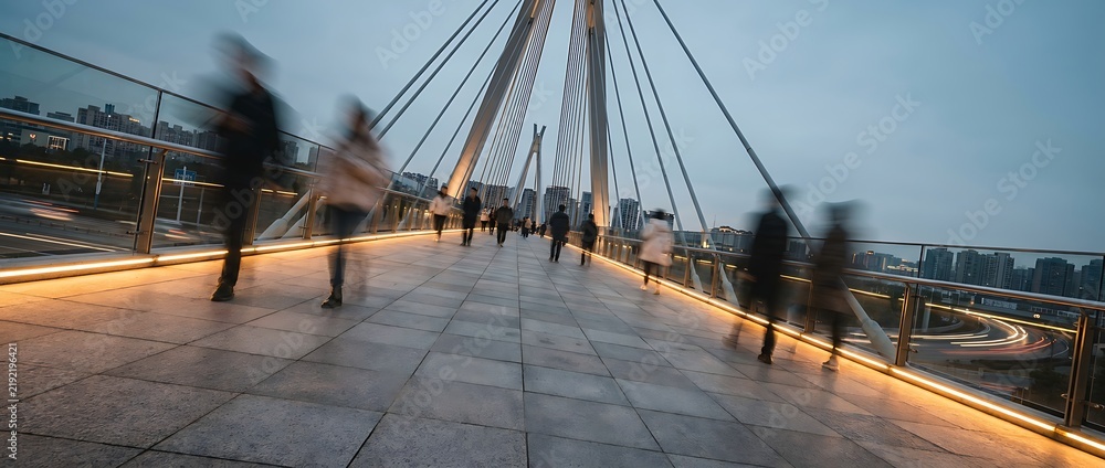 Obraz premium Blurred commuters walking across modern pedestrian cable-stayed bridge during blue hour with city skyline in background and light trails from passing cars creating urban motion and transit concept