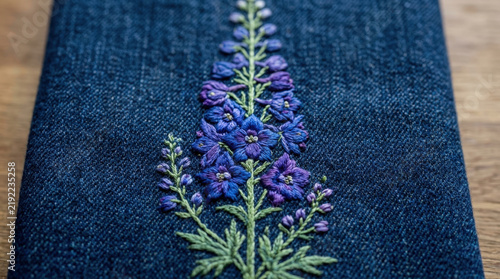 Exquisite blue and purple delphinium flower embroidery on dark blue denim fabric provides elegant botanical decoration for clothing.