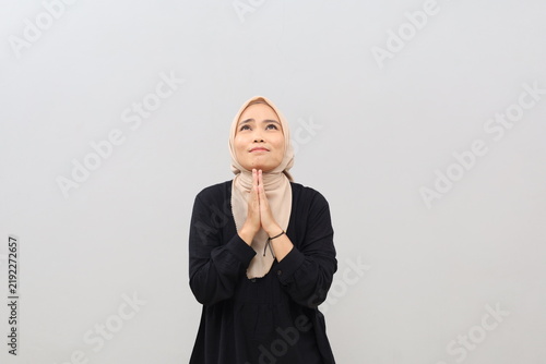 Bild auf Leinwand A young Asian Muslim woman wearing a headscarf displays a hopeful expression, pursing her lower lip and pressing her palms together as if pleading or asking for help, while gazing vacantly upwards