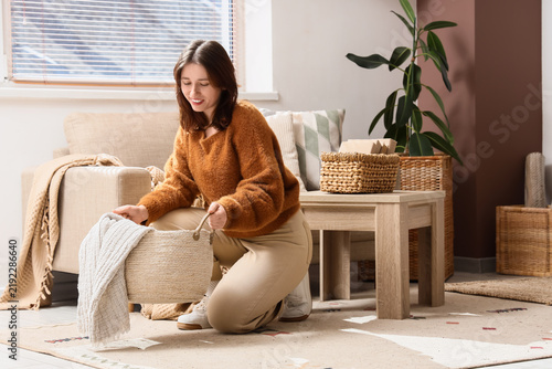 Young woman putting wicker basket with plaid on carpet in living room