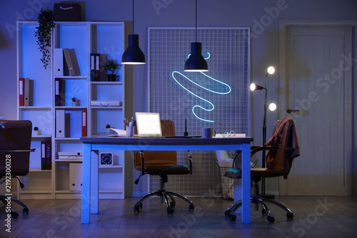 Interior of office with workplace, shelf unit and glowing lamps at night