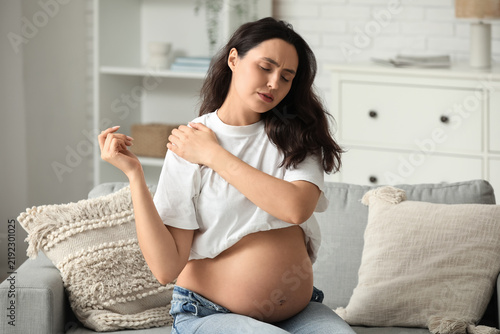 Young pregnant woman suffering from shoulder pain on sofa at home