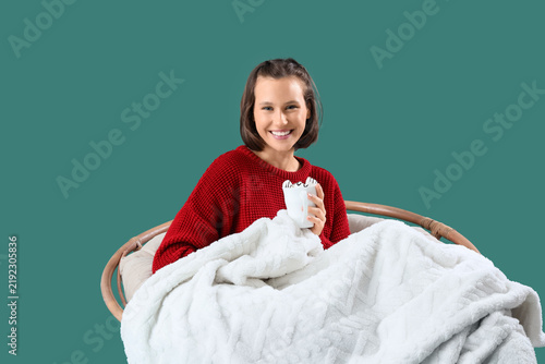 Young woman with warm plaid and cocoa sitting in armchair on green background