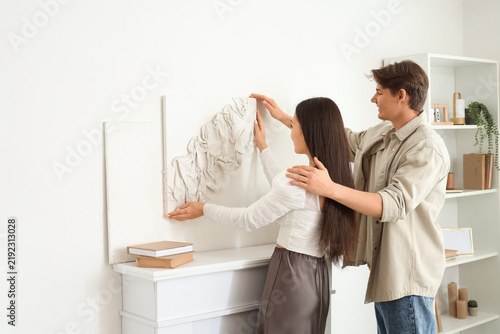 Young couple hanging painting on light wall above fireplace at home