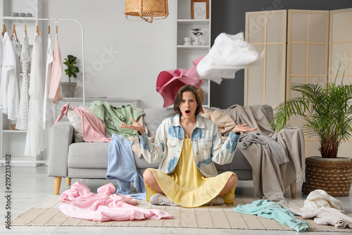 Young disappointed woman sitting on floor with different clothes at home