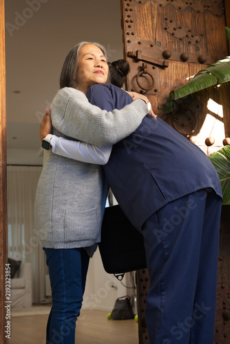 Konstfotografi Senior Asian woman and female caregiver in navy scrubs hugging at doorway with c