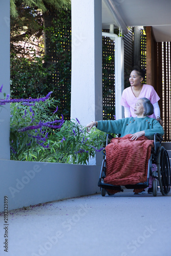 Caregiver in scrubs pushing Senior in wheelchair on patio pointing at planter flowers, copy space