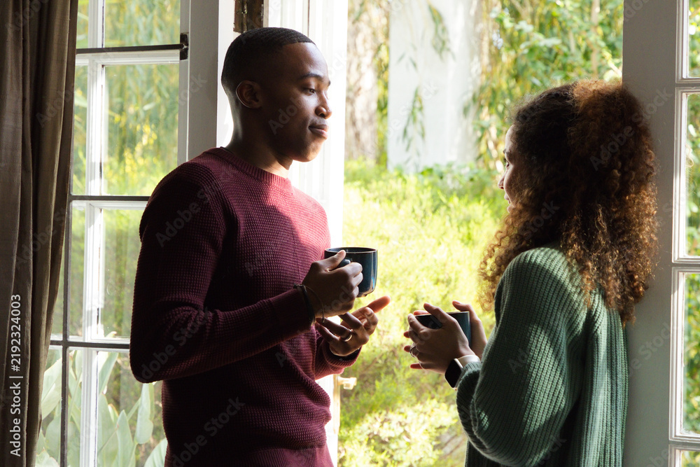 Obraz premium Diverse couple standing by French door, holding mugs, talking with smartwatch visible