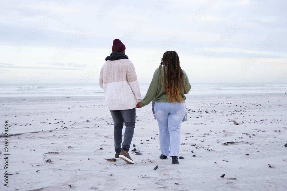 Obraz premium African American couple holding hands walking to sea on beach in knits leaving footprints, pebbles