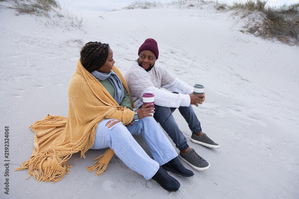 Obraz premium African American couple sitting on sand dune wrapped in mustard blanket, maroon beanie