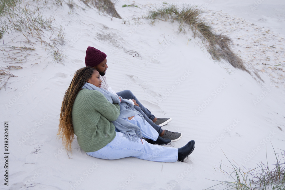 Obraz premium African couple sitting on sand dune at beach wearing maroon beanie and green sweater, copy space