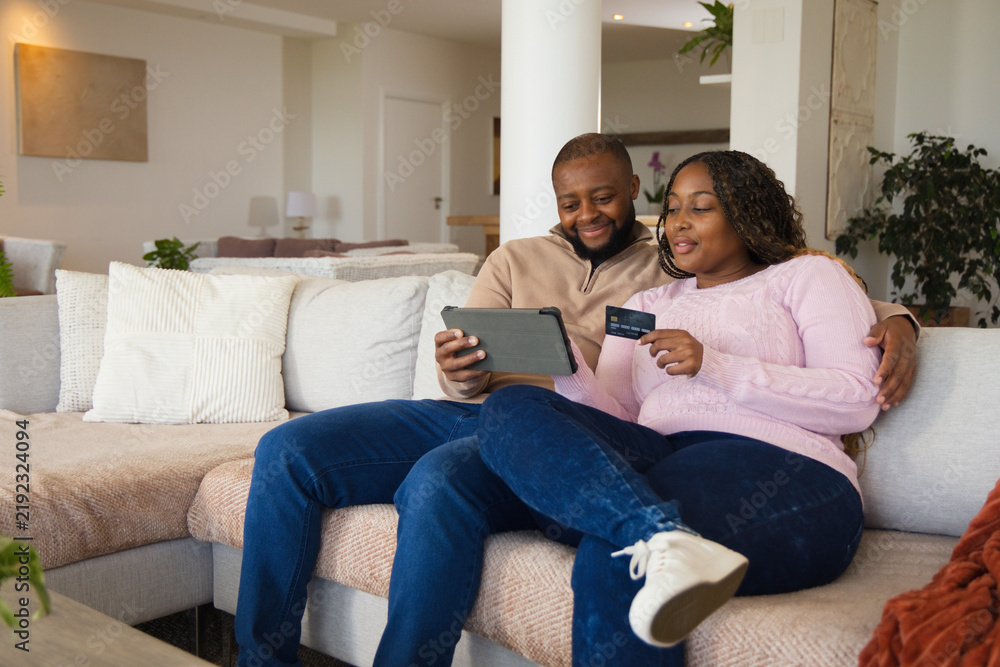 Fototapeta premium African couple sitting on sectional sofa in bright living room, holding tablet and credit card